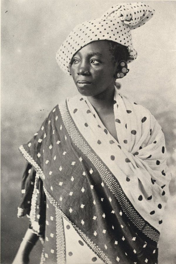 Studio portrait of a Zanzibar woman wrapped in a dark kanga with white spots and a spotted border, with a polka-dot cloth tied on her head