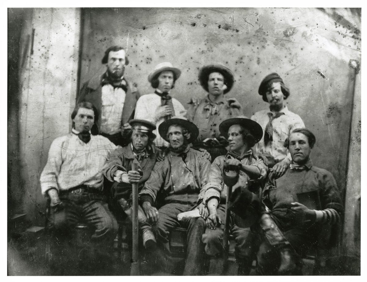 Daguerreotype of nine young men posed as a group in loose work shirts, neckerchiefs, and broad-brimmed hats or caps, two holding shovels