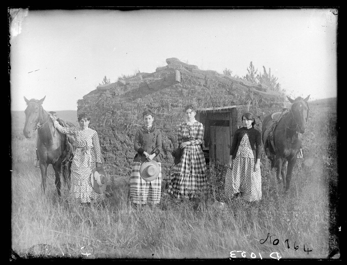 Photograph of four young women standing in front of a sod house with two saddled horses, wearing long dresses in checked and printed cotton fabrics, two holding straw hats