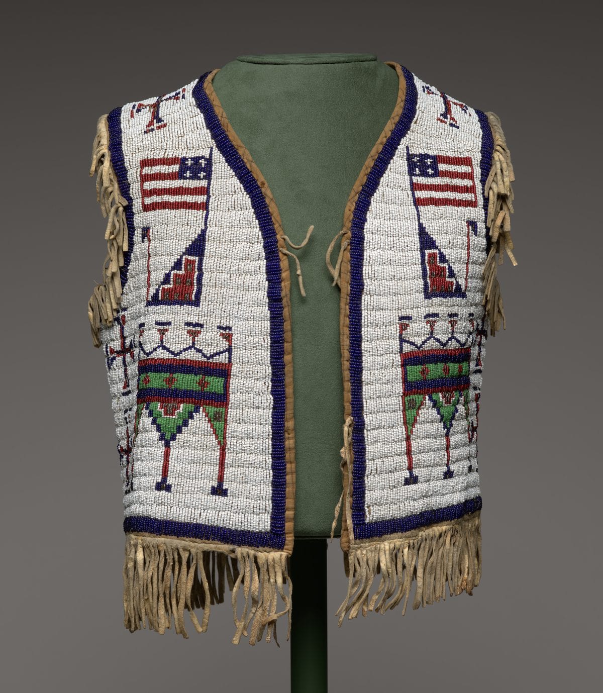 Boy's vest fully covered in white glass beadwork with blue borders, an American flag on each front panel, geometric red and green figures below, and buckskin fringe along the sides and hem
