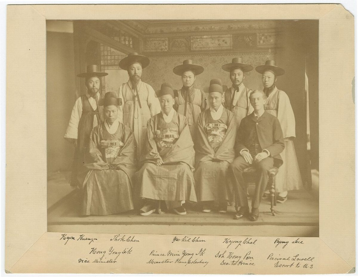 Studio photograph of Korean envoys, three seated in wide-sleeved official robes with embroidered rank badges and five standing in robes and tall black gat hats, beside an American in a Western suit