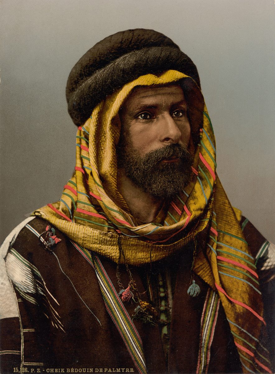 Color photochrom portrait of a bearded Bedouin sheikh in a striped yellow headcloth held by a thick dark headband, and a striped cloak