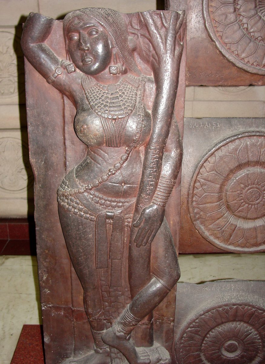 Sandstone railing pillar from Bharhut showing a yakshi grasping a tree, wearing a beaded girdle over a lower garment, a sash with hanging ends, layered necklaces, and heavy anklets