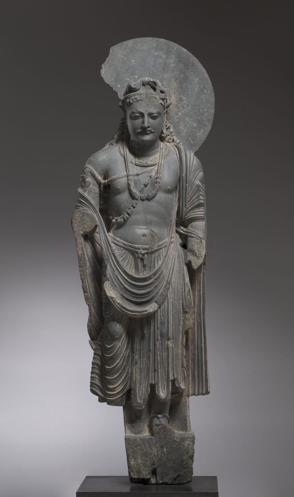 Gray schist statue of a bodhisattva wearing a pleated lower wrap knotted at the waist, an upper cloth looped across the thighs and over the left arm, a jewelled headband, and heavy necklaces