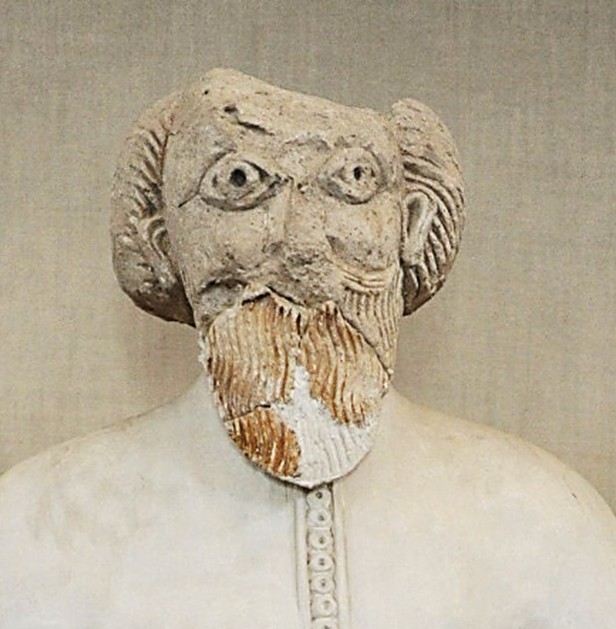 Plaster bust of a bearded man with large staring eyes and curled hair, wearing a smooth garment with a beaded band down the center of the chest