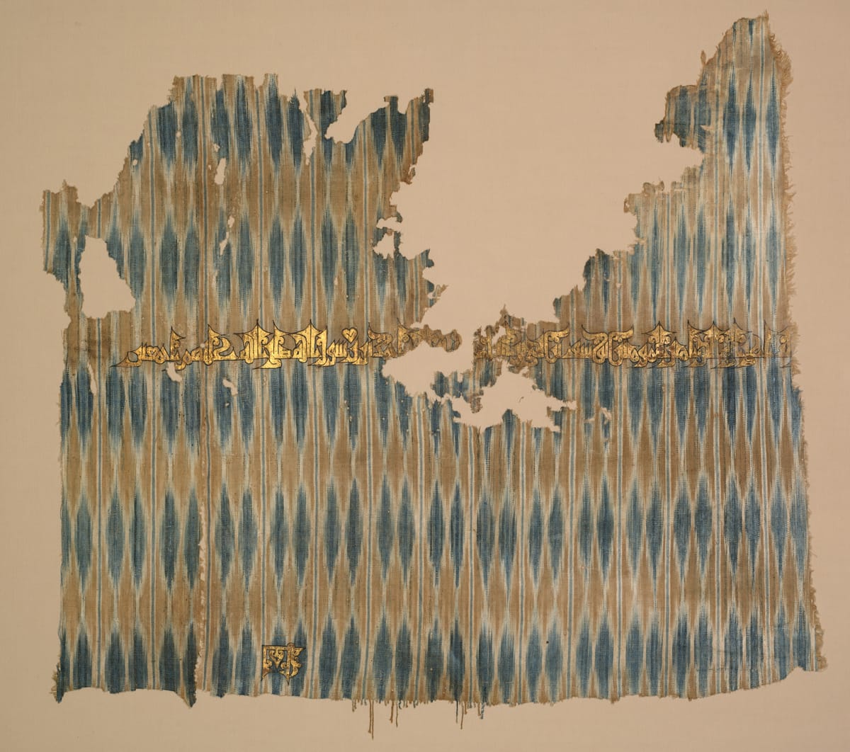 Square of cotton cloth with vertical ikat stripes in blue, brown, and cream, and a band of gold Arabic script across the lower part