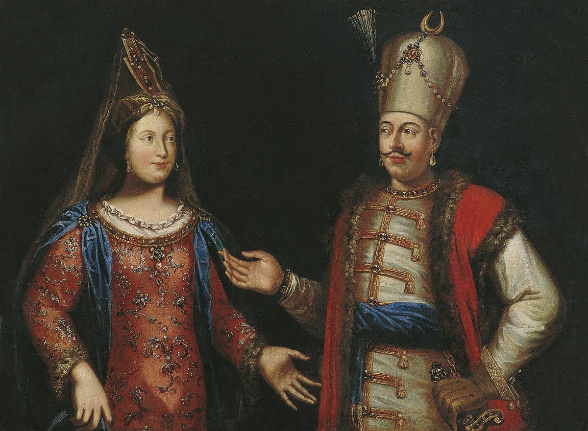 Painting of an Ottoman sultan in a fur-trimmed red kaftan and jeweled tall headdress beside his haseki in an embroidered robe and tall veiled cap