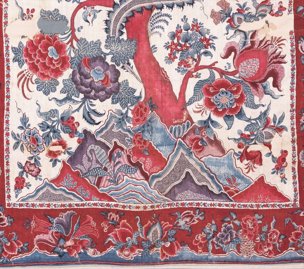Detail of a painted and dyed cotton palampore with a flowering tree rising from stylized red, blue, and purple hills, with a red floral border