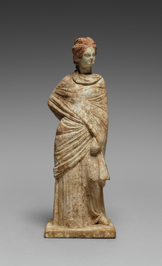 Painted terracotta figurine of a standing woman wrapped in a himation drawn tightly around her body over a long chiton
