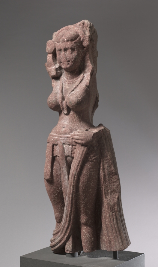 Red sandstone statue of a standing female nature divinity wearing a lower garment held by a girdle at the hips, with long pleated folds falling to the ankles, a heavy necklace, and an elaborate headdress