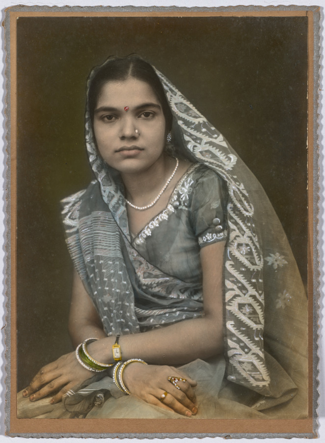 Hand-colored studio photograph of a seated young woman wearing a gray sari with a white patterned border drawn over her head, a short-sleeved blouse, a pearl necklace, and bangles