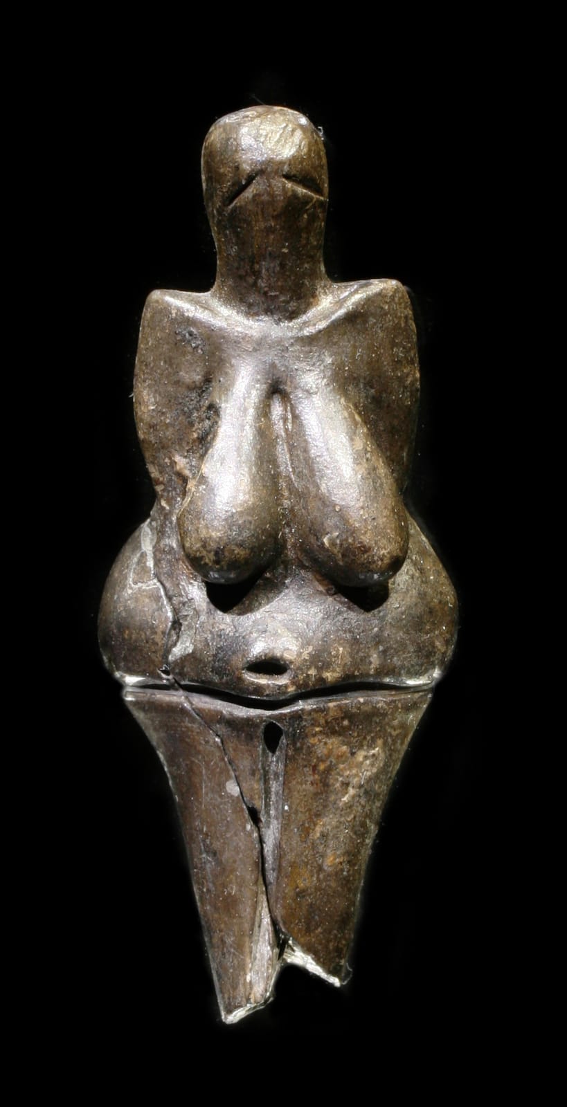 Small dark figurine of a woman with heavy breasts and hips and a simple head, displayed in a museum case