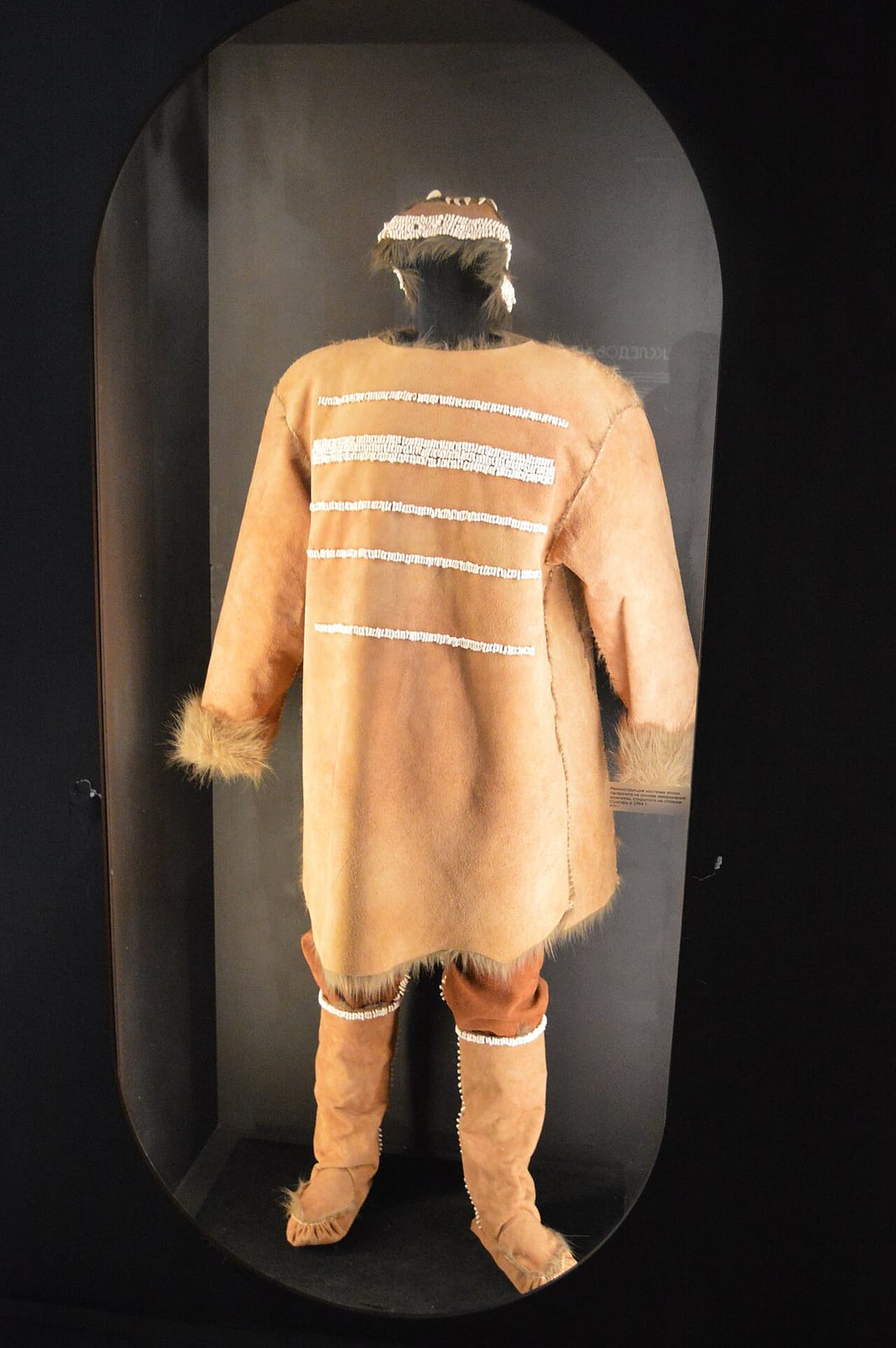 Museum reconstruction of the Sunghir man's clothing seen from the back: a long hide tunic trimmed with fur, rows of white ivory beads across the back, a beaded cap, trousers, and fur-trimmed boots