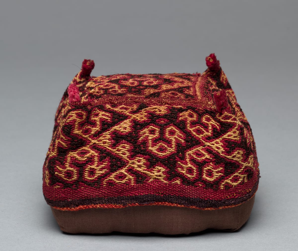 Small square Inca hat with four raised corners and tufted points, covered in a red, gold, and dark brown woven pattern of interlocking birds or animals