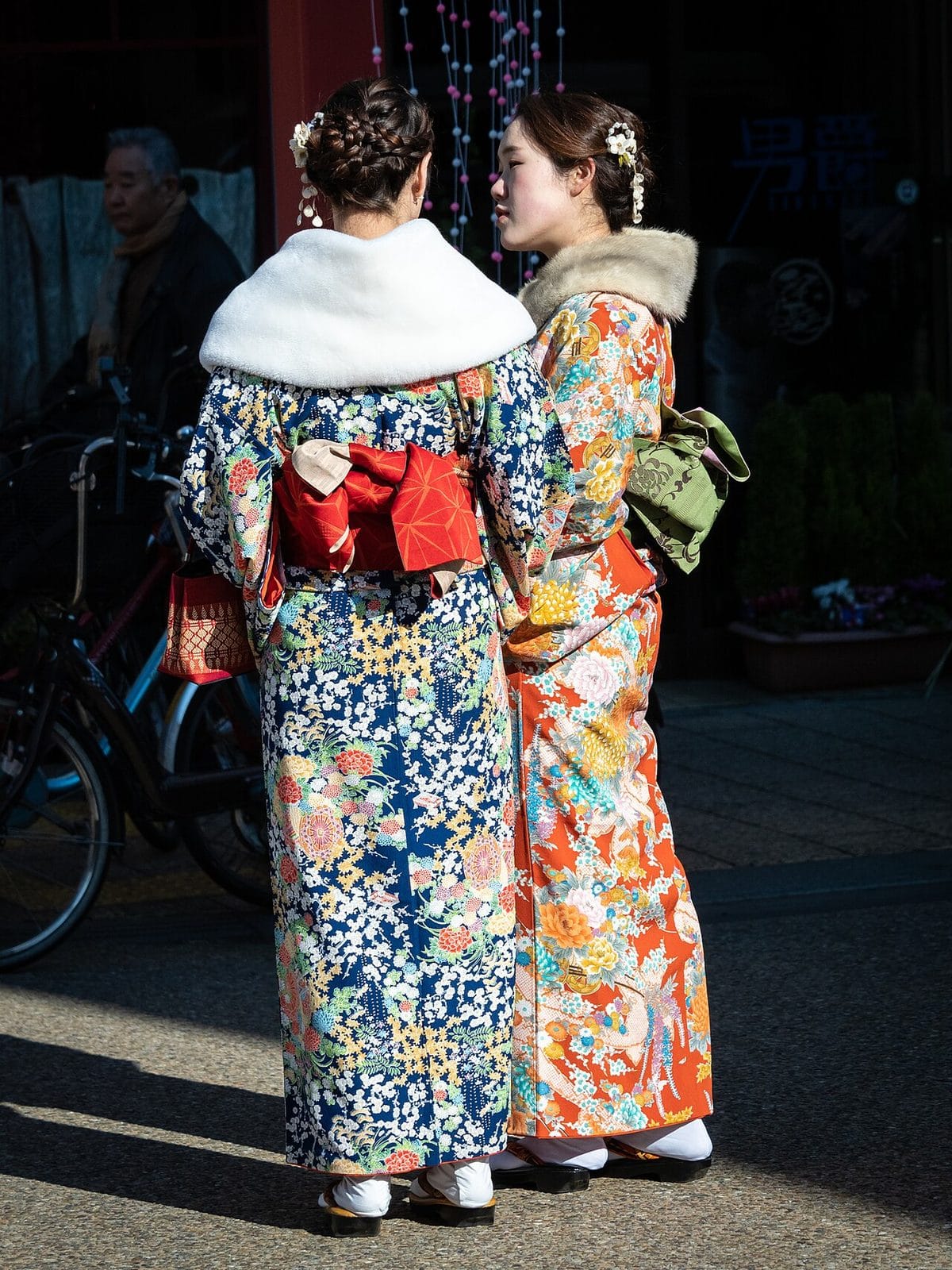 Two young women seen from behind in brightly patterned long-sleeved kimono with tied obi sashes and fur stoles
