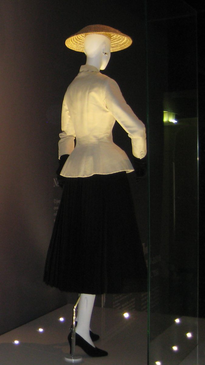 Christian Dior's 1947 Bar suit, a cream fitted jacket with padded hips over a full black pleated skirt, displayed on a mannequin