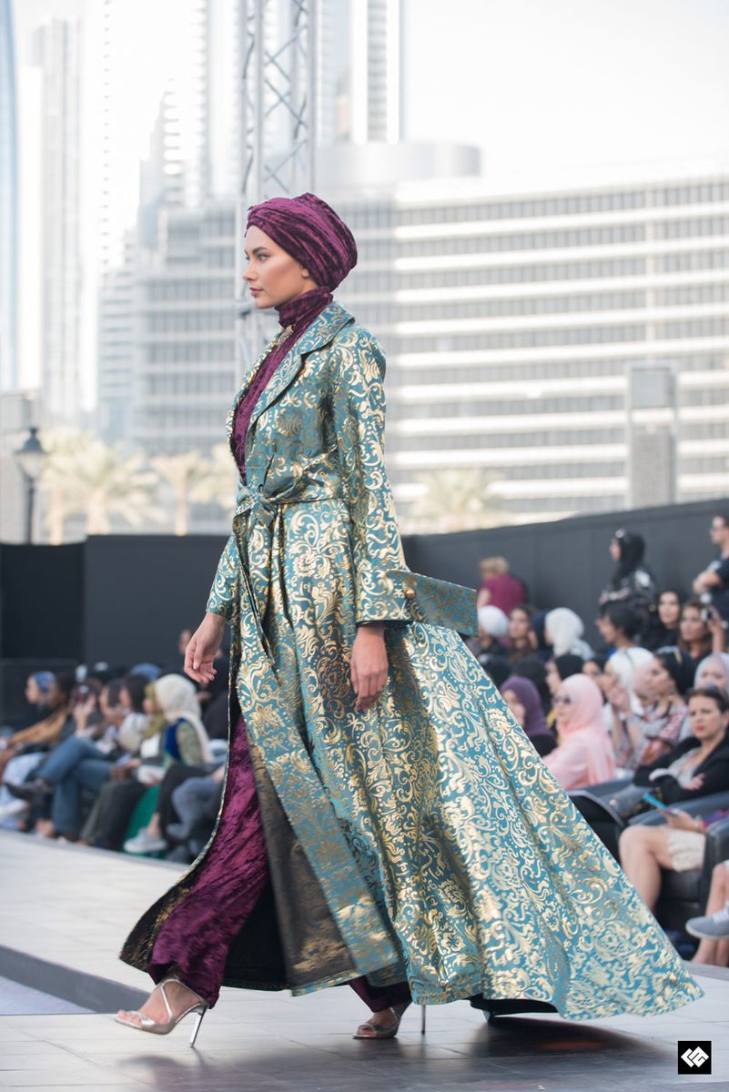 Model on a Dubai runway in a turban-style head wrap and a long teal and gold brocade coat over burgundy velvet