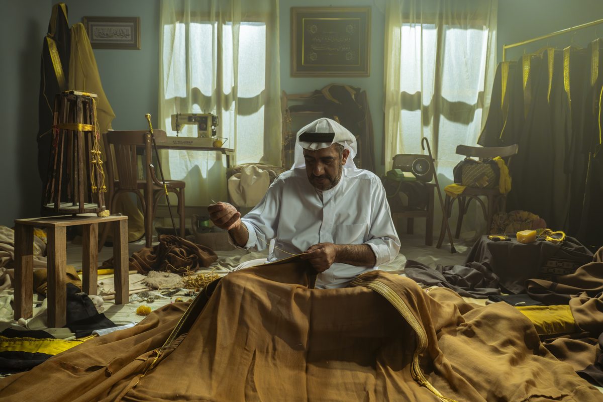 A tailor in a white thobe and ghutra hand-stitching gold trim onto a brown bisht in his workshop in Al-Ahsa, Saudi Arabia