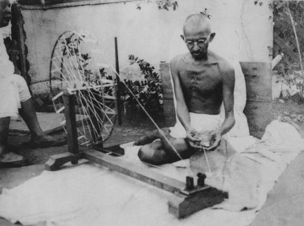 Mahatma Gandhi, bare-chested and wearing a dhoti, seated on the ground spinning thread at a charkha