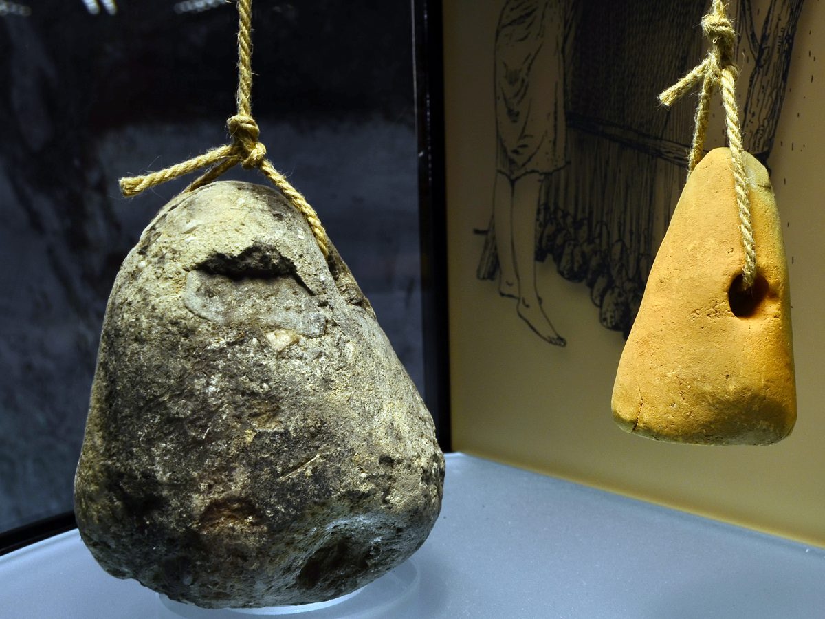 A stone and a clay loom weight hung on cords in front of a drawing of a warp-weighted loom