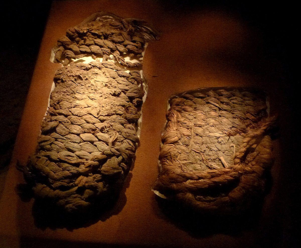 Two worn sandal soles of thick twisted sagebrush-bark cords, twined in tight rows, displayed under a spotlight