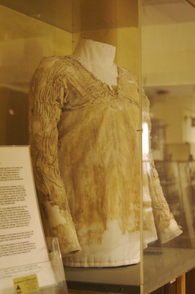 A long-sleeved, finely pleated linen shirt, yellowed and torn at the hem, displayed on a mannequin in a glass case