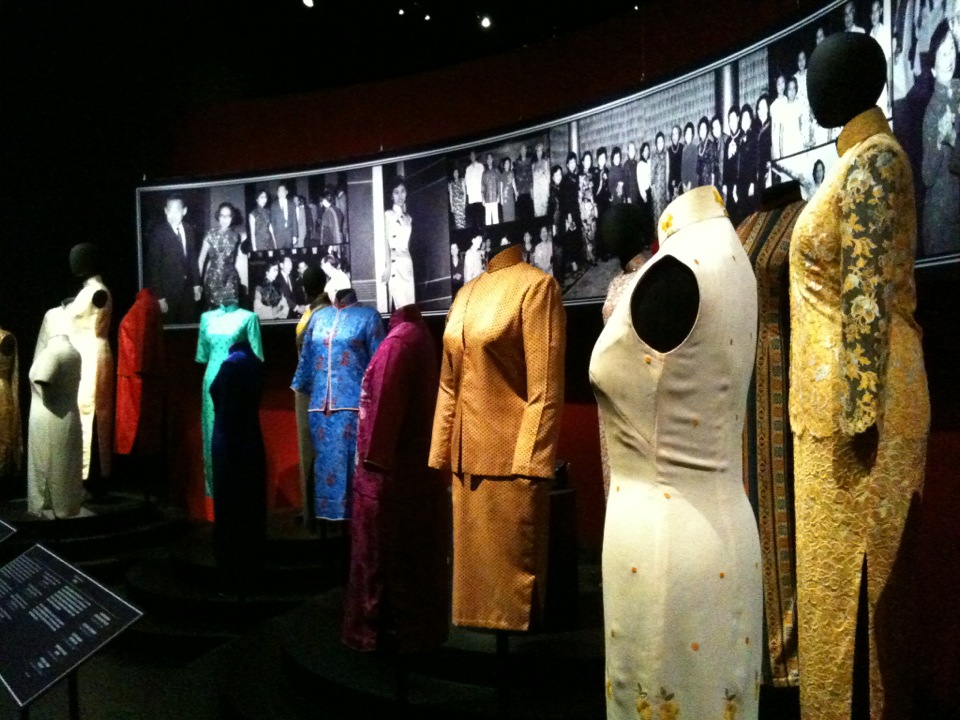 Mannequins in a dark gallery wearing cheongsam in white, gold lace, turquoise, and blue, in front of a wall of black-and-white photographs