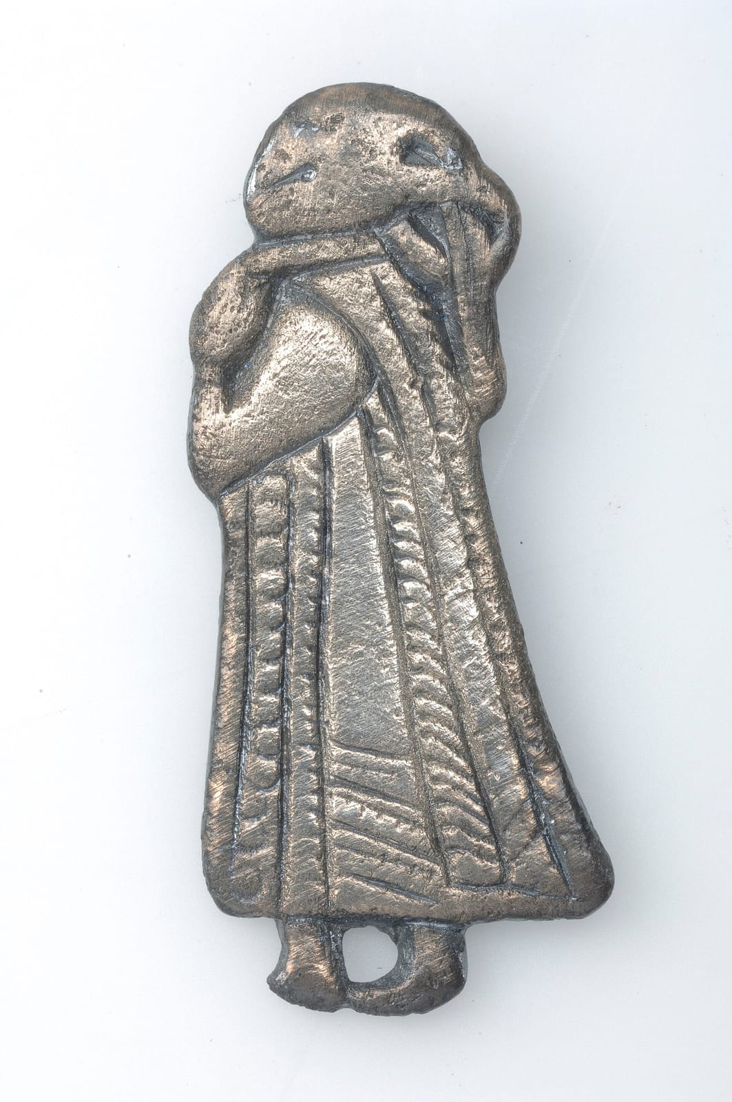 Silver pendant of a standing female figure in a long garment with vertical bands of decoration, a trailing hem, and hair tied in a knot at the back